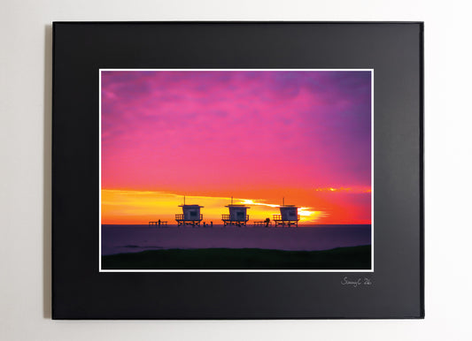 Three lifeguard towers on Santa Monica Beach under a pink and yellow winter sunset