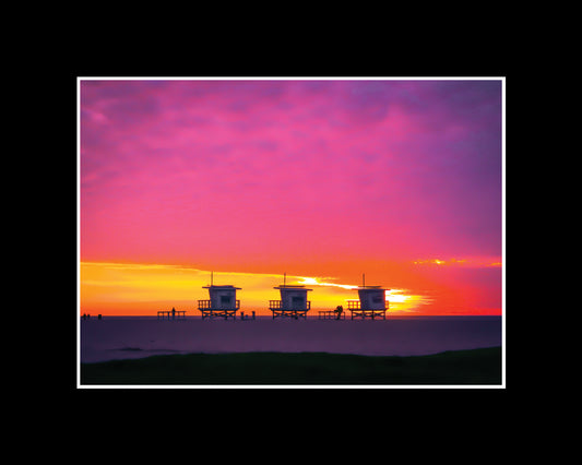 Three lifeguard towers on Santa Monica Beach under a pink and yellow winter sunset